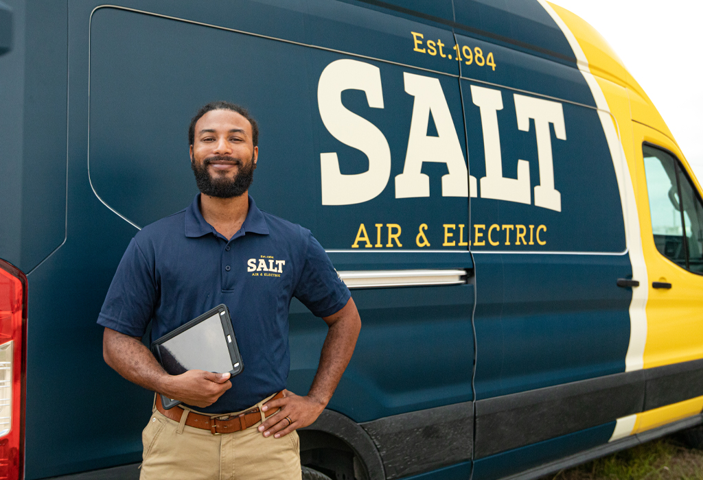 SALT technician in a navy uniform holding a tablet, standing professionally in front of a branded company service van featuring a yellow and navy design