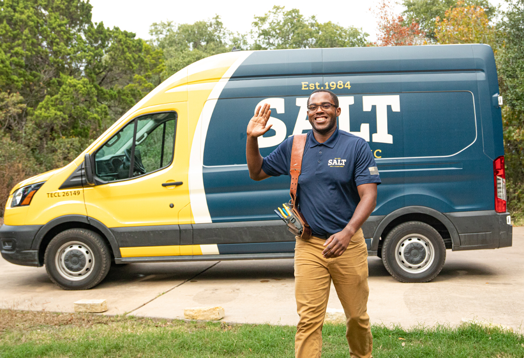 SALT technician in navy uniform waving while walking in front of a yellow and navy branded company service van parked on a driveway