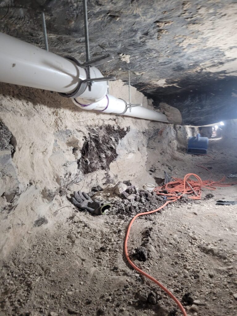 PVC plumbing pipes installed in a crawl space with visible dirt, rocks, gloves, and orange extension cord