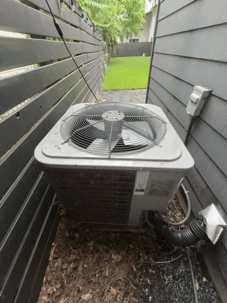 Outdoor air conditioning condenser unit installed in a narrow side yard between a house and wooden fence, with leaves and grass visible around the base.