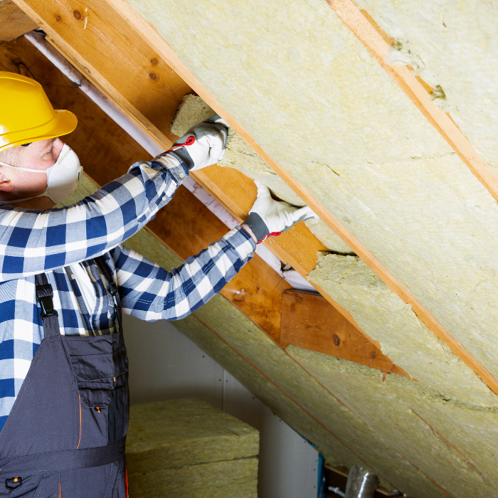 Worker wearing a hard hat and protective mask installing insulation between wooden rafters inside an attic space.