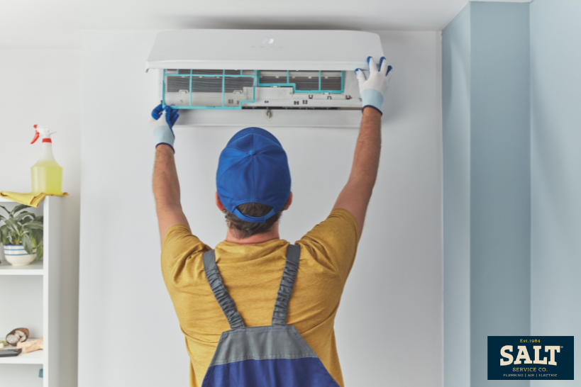 HVAC technician installing or servicing a wall-mounted mini split air conditioner, lifting the front panel while wearing gloves and work overalls.