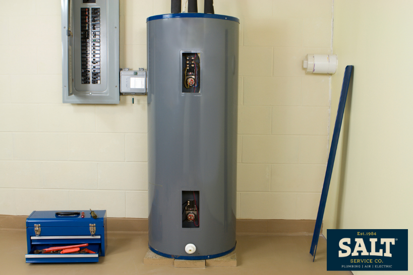 Plumber installing or servicing a wall-mounted water heater in a bathroom, wearing a tool belt and cap during maintenance.