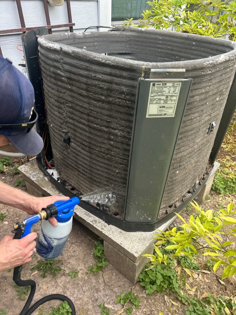 Technician cleaning a dirty outdoor AC condenser coil during HVAC maintenance.