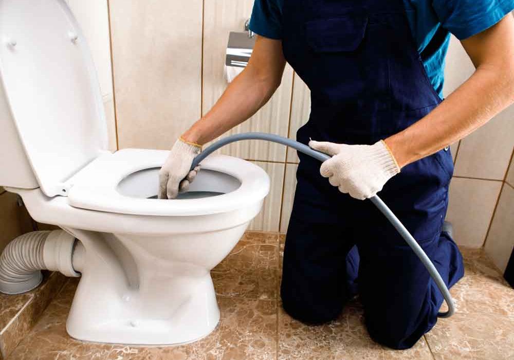Plumber in navy overalls kneeling on a tiled bathroom floor, using a drain snake tool to unclog a residential toilet