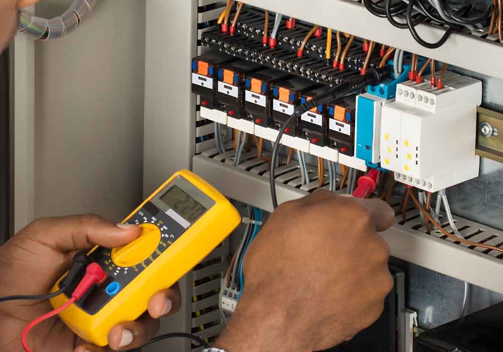 Electrician using a yellow multimeter to test voltage in an open electrical panel with multiple wires and circuit breakers