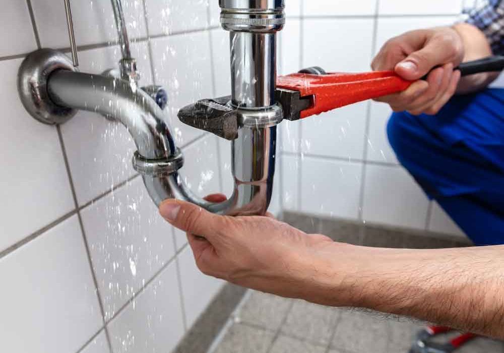 Plumber using a red pipe wrench to tighten a leaking chrome drainpipe under a tiled sink with water spraying from the joint